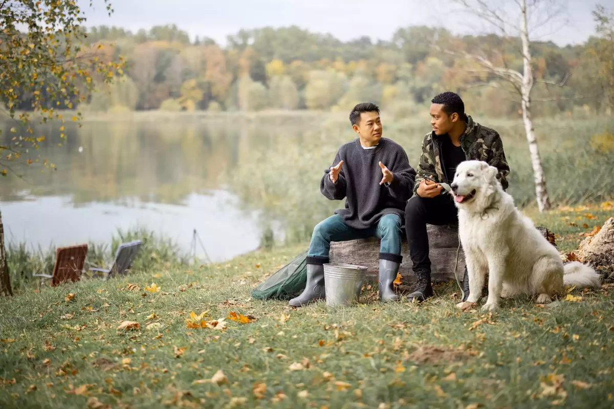 Two male friends talking with dog sat beside them next to a river.